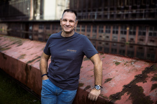 Zu sehen ist ein Mitarbeiter der Firma Gelsenbad. Er trägt ein Firmenhemd und steht lässig mit freundlichen Lächeln an einem Industriegebäude.